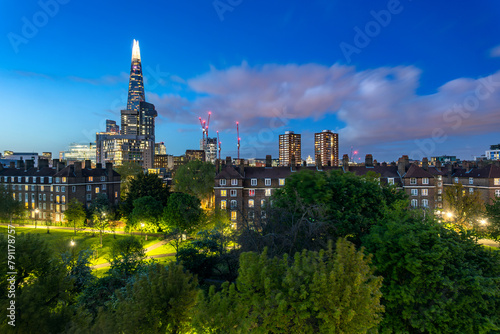 A residental neighbourhood with a geen park and illuminated buildings including skyscrapper Shard in London during blue hour