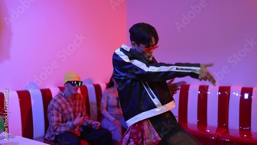 Asian man standing and dancing with pop music while multicultural friend sitting behind at home with led light. Attractive street dancer moving to hip hop music while looking at camera. Regalement. photo