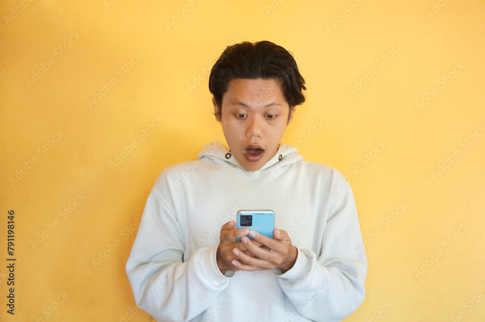 Fototapeta premium Surprised Asian young man wearing a hoodie while holding and looking at mobile phone