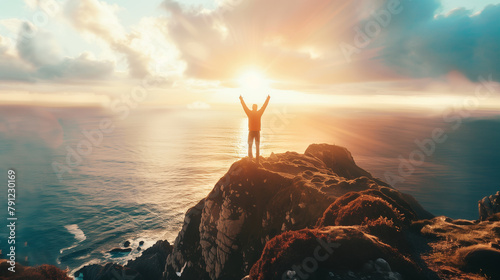 A man stands on a hill overlooking the ocean, with the sun shining brightly on him. He is holding his arms up in the air as if he is celebrating or expressing joy. Concept of freedom and happiness
