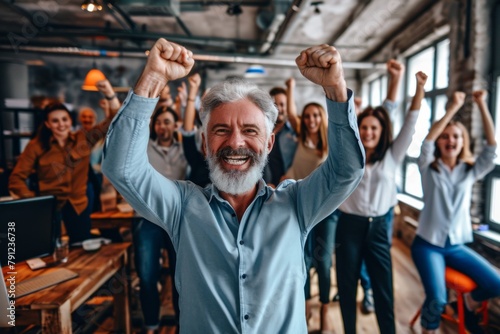 Portrait of a happy senior businessman raising his hands with his team in the background. © Loli