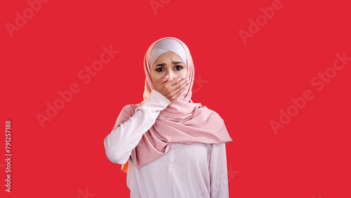 Secret fear. Shocked face. Dismay panic. Scared disturbed upset woman in headscarf covering mouth with hand isolated on red empty space background.