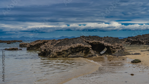 Low tide. Many attached mollusks can be seen on the exposed rocks. The waves of the ocean foam and spread on the sandy shore. Clouds in the sky. Madagascar. Nosy Tanikely