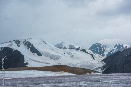 Dramatic vast misty top view from ice pass to big glacier tongue among sharp rocks and large snow-capped mountains in rainy low clouds. Dark atmospheric mountain silhouettes in rain in gray cloudy sky