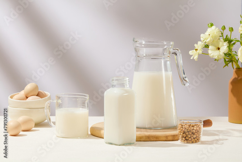 A stack of bowl containing eggs displayed with some glass containers filled with milk and a cup of soybeans. A pot of flowers featured over white background. Healthy breakfast promotion