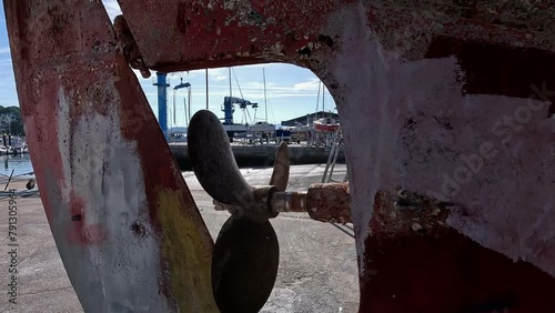 The rusty engine and rudder propeller of the old fishing boat in the shipyard for restoration with the harbor cranes in the background, close-up shot traveling backwards. Portonovo, Galicia, Spain photo