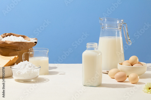 White table features several types of glass containers filled with milk, bowls of eggs and sandwiches and flour inside a paper bag. Empty space for product presentation with baking content