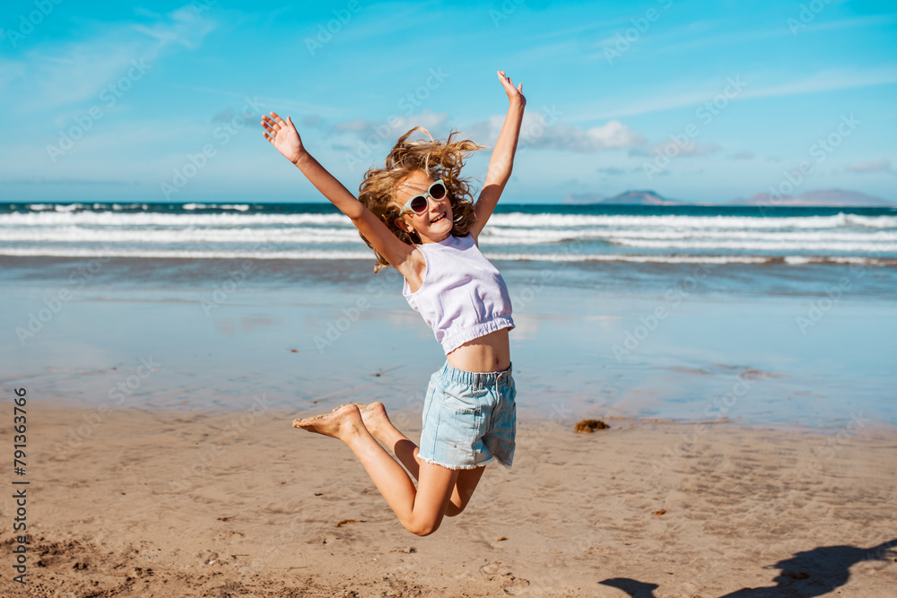 Jumping girl on beach. Smilling blonde girl enjoying sandy beach, looking at crystalline sea in Canary Islands. Concept of beach summer vacation with kids. - obrazy, fototapety, plakaty 