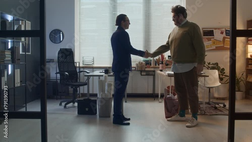 Full shot of two feuding office colleagues getting up from desks, walking towards each other, funny guy giving man in suit handshake with wooden toy hand, jumping and dancing with joyFull shot of two  photo