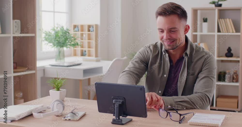 Happy young man, sitting at his table at home, uses a modern digital tablet with a stand, makes a video call, says hi, takes off his glasses, talks to his friend, shares news, and has lots of fun Happy young man, sitting at his table at home, uses a modern digital tablet with a stand, makes a video call, says hi, takes off his glasses, talks to his friend, shares news, and has lots of fun