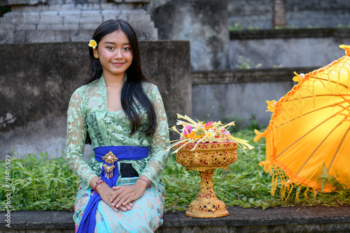 Young woman in traditional Balinese attire smiling beside a vibrant yellow umbrella and a decorative golden basket. 