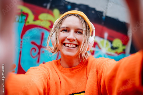 Happy young woman wearing wireless headphones and taking selfie in front of graffiti wall photo