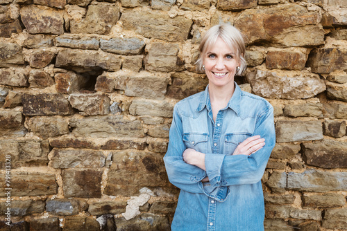 Confident blonde woman with short hair and denim shirt, smiling outdoors with crossed arms in front of a stone wall