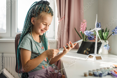 Smiling girl plugging smart phone near wind turbine model at table photo