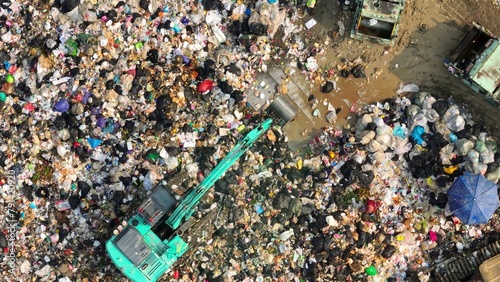 A landfill sprawls, a mosaic of refuse. A teal excavator toils beside a truck, amidst the chaos of mixed waste. The scene echoes humanity's consumption and the tireless effort of waste management.  

