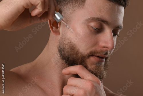 Handsome man applying serum onto his face on brown background