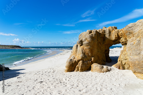 Arche naturelle sculptée dans la roche, encadrant une plage de sable blanc et des eaux turquoises, sous un ciel bleu, en Finistère Nord, Bretagne.