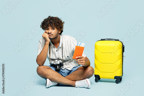 Full body sad traveler Indian man wear white casual clothes sit hold bag passport ticket isolated on plain blue background. Tourist travel abroad in free time rest getaway. Air flight journey concept. #791448593