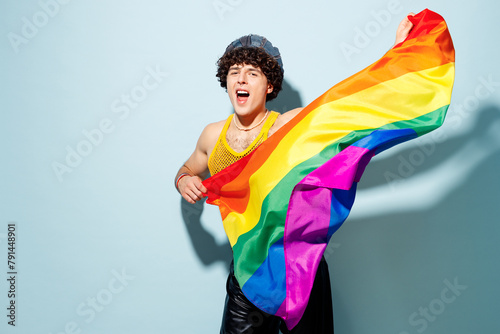 Young fun happy gay Latin man wear mesh tank top hat clothes hold striped rainbow flag sing isolated on plain pastel light blue cyan background studio portrait. Pride day June month love LGBT concept.