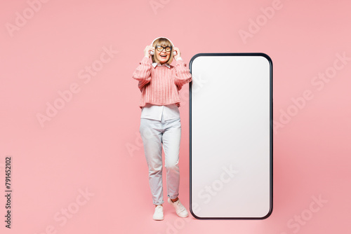 Full body elderly woman 50s year old in sweater shirt casual clothes glasses big huge blank screen mobile cell phone smartphone with area listen music in headphones isolated on plain pink background