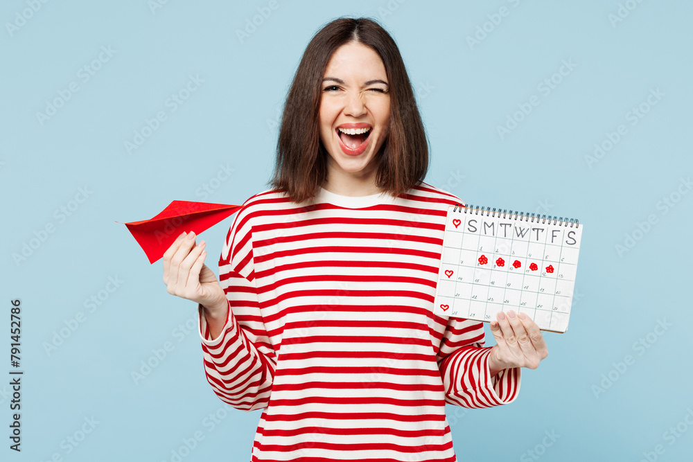 Young excited surprised woman wear red casual clothes hold paper airplane female periods pms calendar checking menstruation days wink isolated on plain blue background. Medical gynecological concept. Young excited surprised woman wear red casual clothes hold paper airplane female periods pms calendar checking menstruation days wink isolated on plain blue background. Medical gynecological concept.
