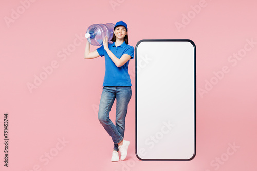 Full body happy delivery employee woman wears blue cap t-shirt uniform workwear work as dealer courier big huge blank screen area mobile cell phone hold water bottle isolated on plain pink background.