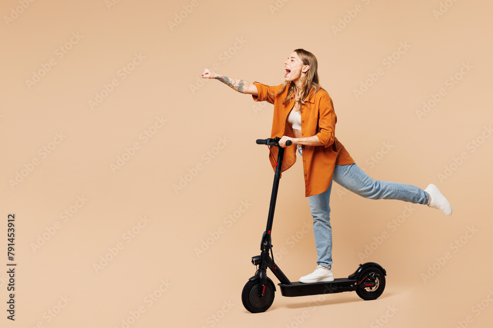 Full body side view young woman wear orange shirt casual clothes ride electric scooter do super hero power gesture isolated on plain pastel light beige background studio portrait. Lifestyle concept. - obrazy, fototapety, plakaty 