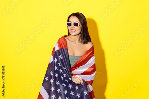 A stylish woman in sunglasses in a swimsuit holds an American flag on a yellow background. The concept of recreation in the USA