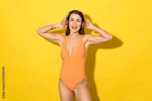 An attractive tanned woman in a yellow swimsuit listens to music on a yellow background. Summer holidays, moods of joy and sunshine
