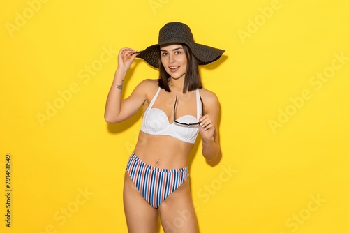 An attractive tanned woman in a swimsuit poses on a yellow background. A bright concept of summer holidays