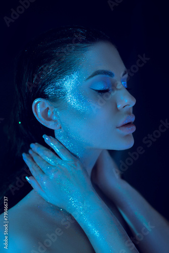 Woman with shimmering blue cosmetic makeup, serene pose, face and hand glowing under a soft blue light