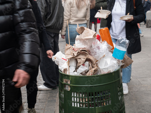 overflowing garbage bin in the city with blurred and unrecognizable people walking in the background