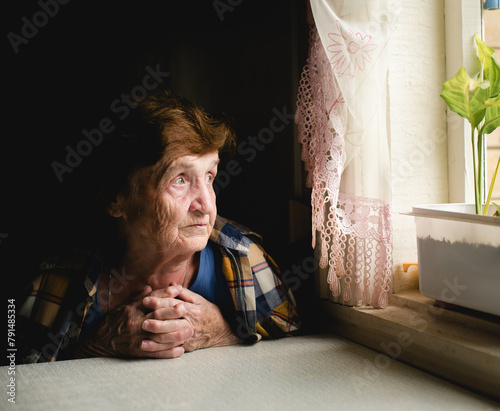 An elderly woman sits by the window, gazing outside with a thoughtful expression, lost in contemplation.