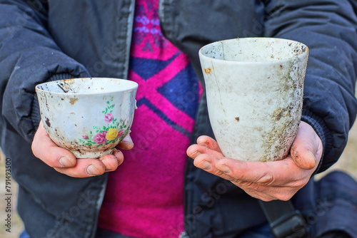 Fototapeta Naklejka Na Ścianę i Meble -  two old  dirty broken different white empty ceramic large and small tea cups in the hands of a man dressed in a black jacket on the street during the day