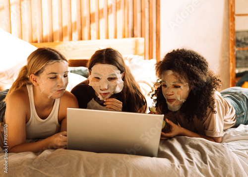 Charming mixed-race teenagers in cosmetic fabric masks on their faces are looking at laptop. Multiethnic friends have fun and use computer.