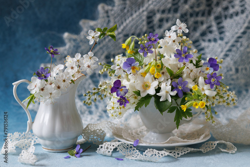 Beautiful still life with spring flowers, a bouquet of white and blue flowers in a cup and jug on a decorative background with a white shawl.