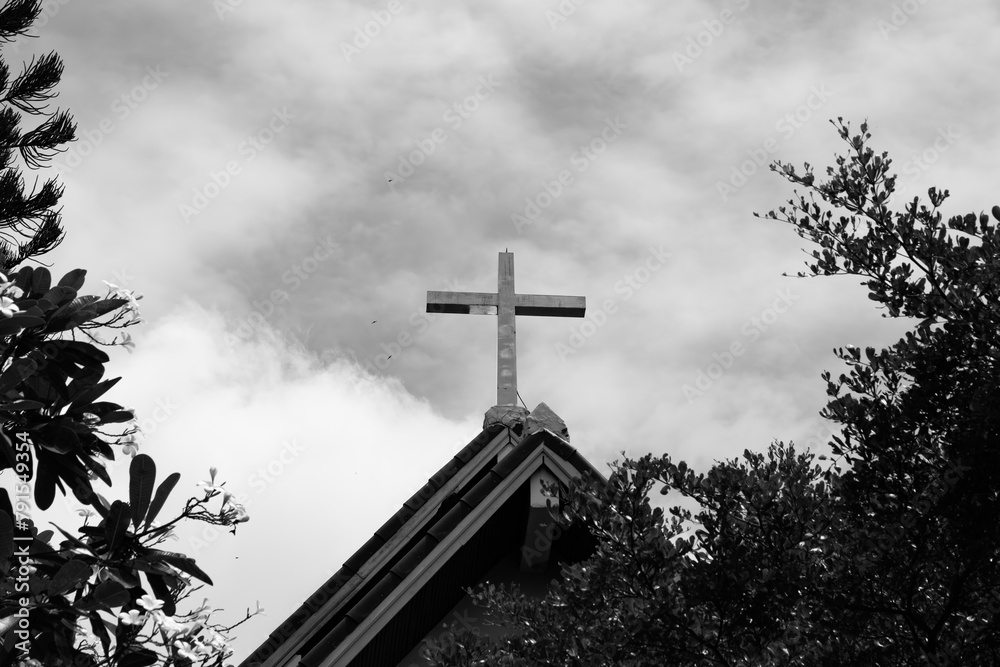 A cross is on top of a roof in front of a cloudy sky. The cross is the only thing visible