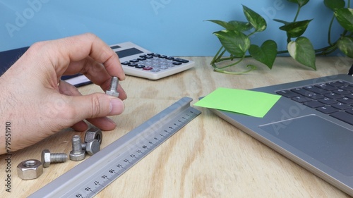Man's hand holds a screw, on desk in engineering office with compiuter and ruler.