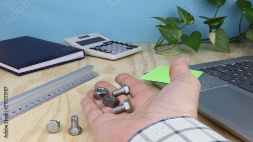 Man's hand holds group of screws and bolts, on office desk with agenda, computer and ruler.