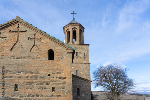 Part of the church and bell tower of the Shavnabada monastery. Stone yard. Brick walls. Tree without leaves. Bright blue sky with clouds. Georgia.