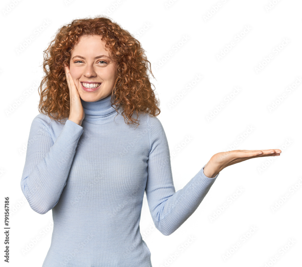 Young Caucasian redhead woman holds copy space on a palm, keep hand over cheek. Amazed and delighted. Young Caucasian redhead woman holds copy space on a palm, keep hand over cheek. Amazed and delighted.
