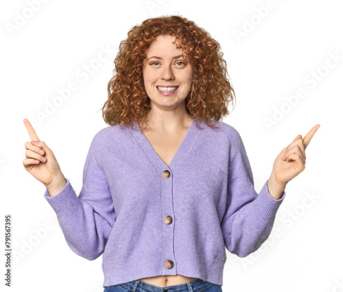 Young Caucasian redhead woman indicates with both fore fingers up showing a blank space.