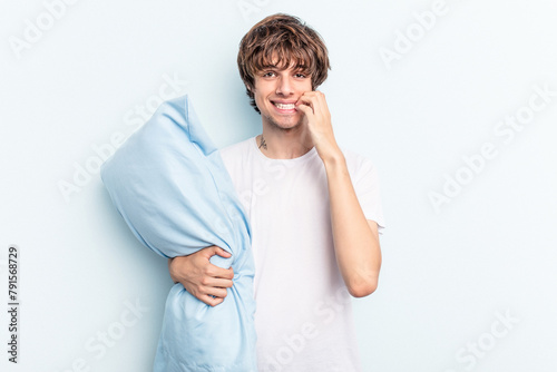 Young caucasian man going to sleep holding a pillow isolated on blue background biting fingernails, nervous and very anxious.