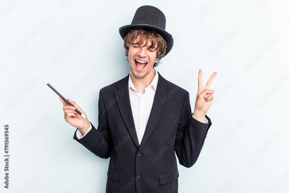 Young magician man holding magic cards isolated on blue background joyful and carefree showing a peace symbol with fingers. Young magician man holding magic cards isolated on blue background joyful and carefree showing a peace symbol with fingers.