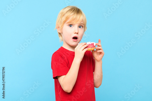 Cute little boy with blond hair and freckles eating a glazed donut. Children and sugary junk food concept. Boy holding colorful donuts, eating junk unhealthy food full of sugar.