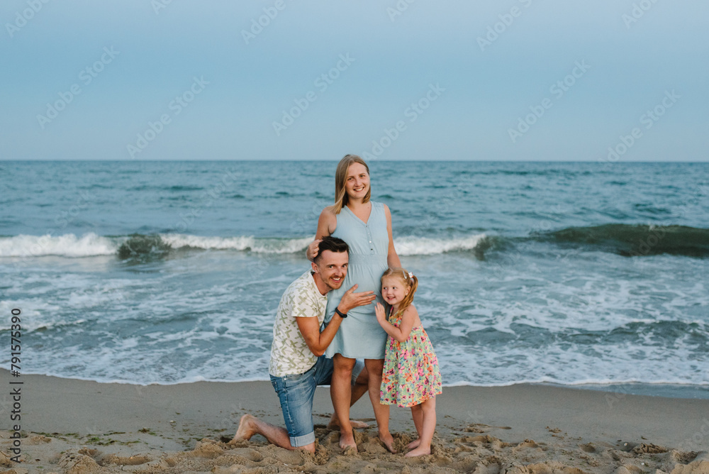 Naklejka premium Mother, father and child in love hugging on seashore. Happy family hugs a child barefoot standing on a sand beach. Man hugs pregnant wife. Dad, daughter kisses stomach mom enjoy summer vacation on sea