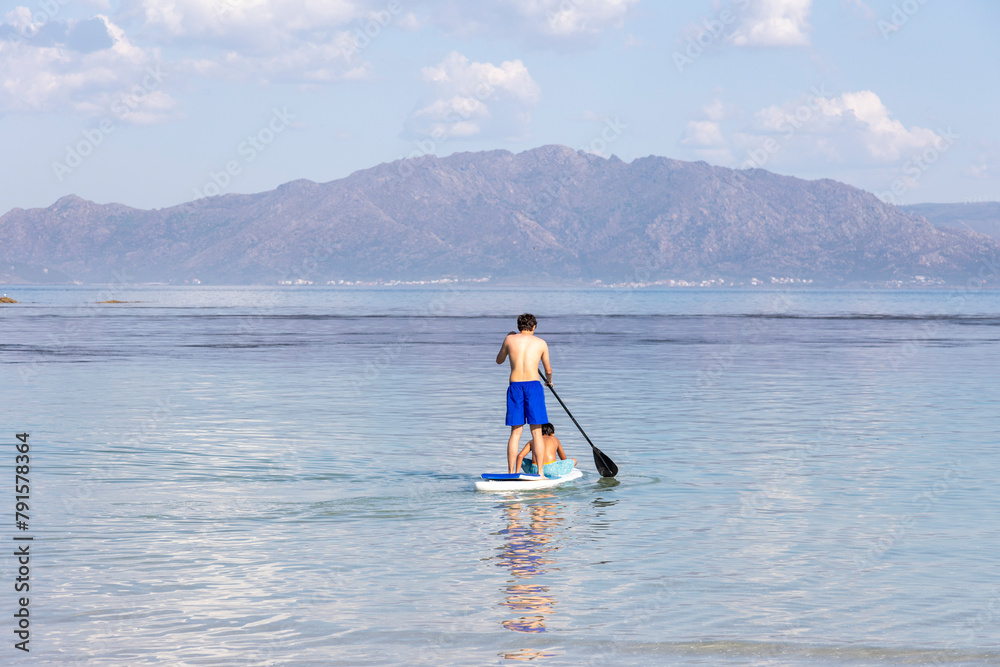 Naklejka premium A lone man enjoys paddleboarding on the tranquil blue waters with a mountainous backdrop, reflecting serenity and the simplicity of nature