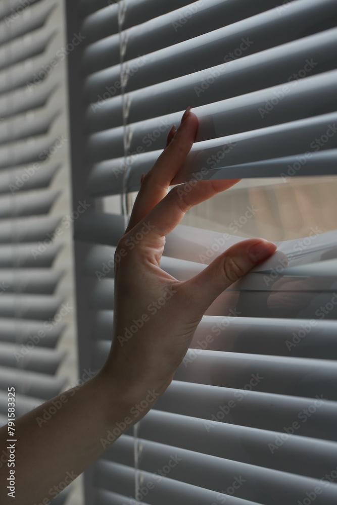 Naklejka premium Woman separating slats of white blinds indoors, closeup