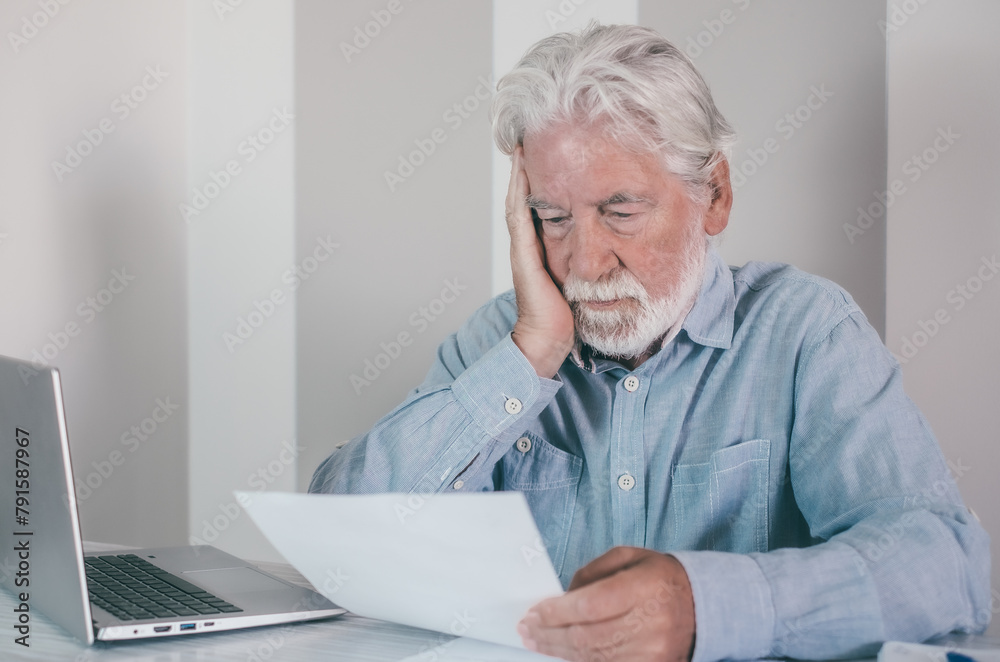 Serious bearded senior man sitting at home check the results of his medical tests before to talk remotely with doctor on laptop webcam to have doctor's consultation Serious bearded senior man sitting at home check the results of his medical tests before to talk remotely with doctor on laptop webcam to have doctor's consultation