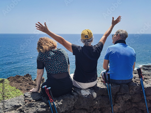 Back view of three friends in outdoor excursion at sea, sitting on the cliff looking at the horizon over water, enjoying freedom and vacation, healthy activity in retirement